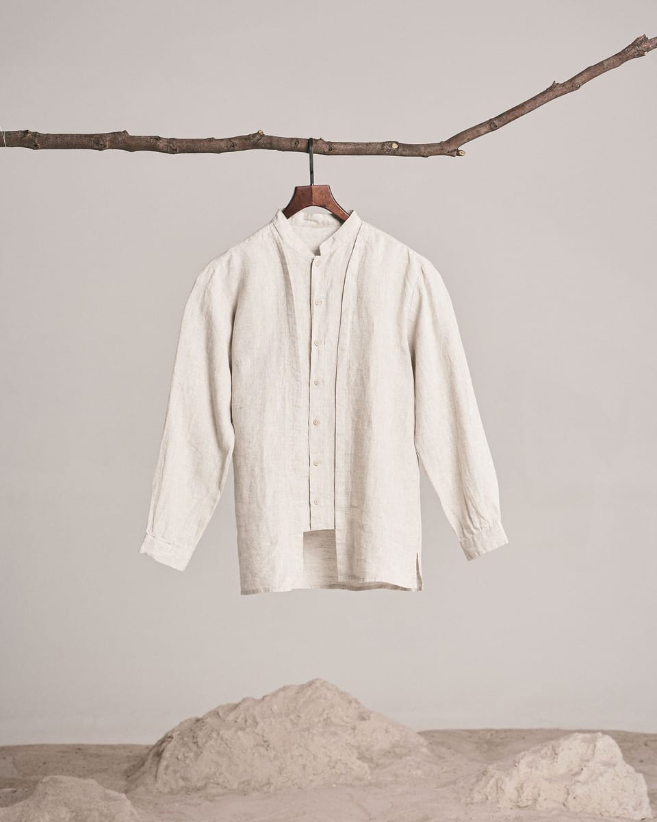 La camisa Ponent de lino crudo con cuello mao, colgada de una percha en una rama